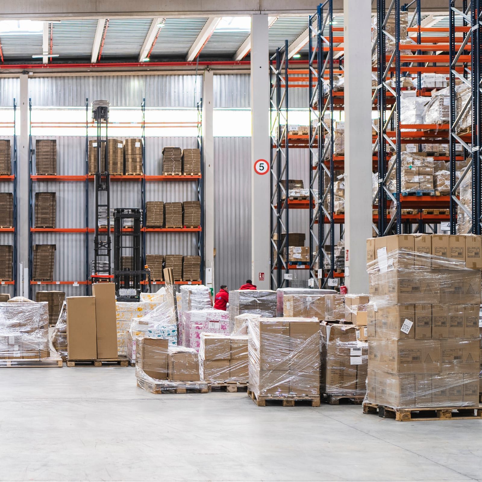 Pallets met gewikkelde dozen die los op de vloer van een magazijnhal staan, met daarachter stellingen met genummerde locaties.