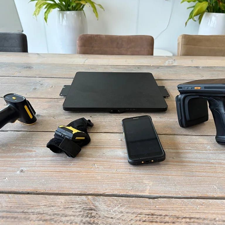 Vijf leesapparaten op een houten tafel: een pistoolscanner, een draagbare scanner met band, een plat leesplateau, een robuuste telefoon en een RFID-handheld met pistoolgreep.